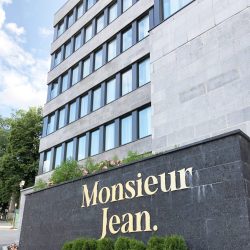 Façade moderne de l’Hôtel Monsieur Jean à Québec avec enseigne dorée, bâtiment en pierre grise et aménagement paysager fleuri à l’entrée