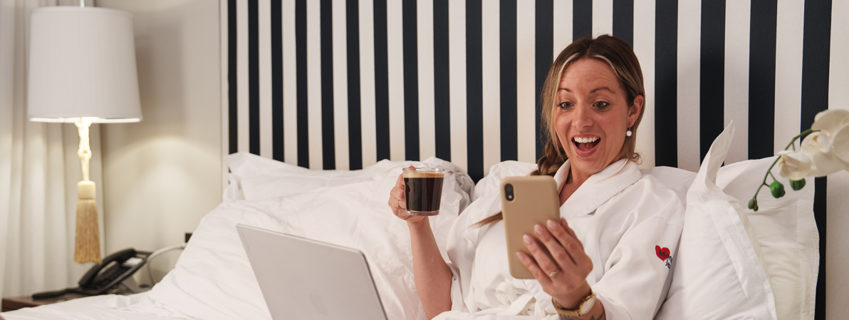 Femme en peignoir buvant un café au lit et utilisant son téléphone dans une chambre d’hôtel élégante