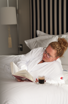 Personne détendue lisant un livre dans un lit d’hôtel confortable, vêtue d’un peignoir blanc avec un café à proximité, dans une ambiance calme et reposante.