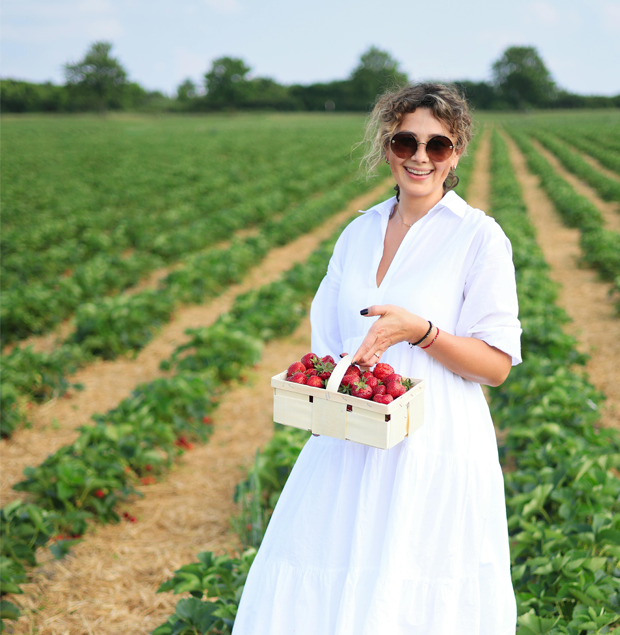 Femme souriante tenant un panier de fraises fraîches dans un champ cultivé, entourée de rangées de plants sous un ciel dégagé.