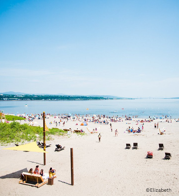 Vue panoramique de la Baie de Beauport avec une grande plage animée, des visiteurs profitant du bord de l’eau et du ciel dégagé en arrière-plan.