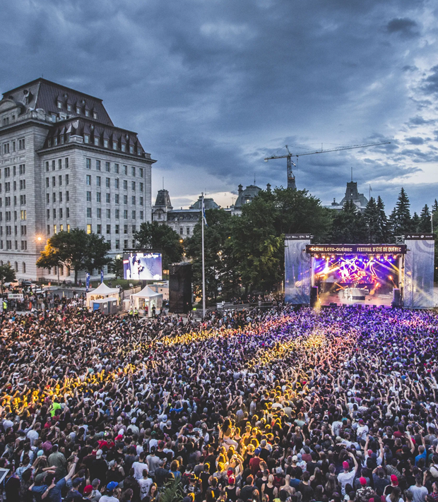Foule rassemblée devant une grande scène extérieure lors du Festival d’été de Québec, dans une ambiance festive sous un ciel orageux en soirée.