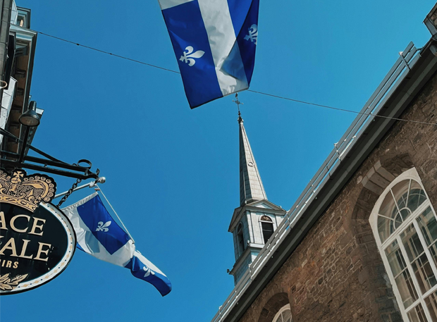 Drapeaux du Québec flottant dans le Vieux-Québec devant une église historique en pierre, sous un ciel bleu éclatant lors de la saison estivale.