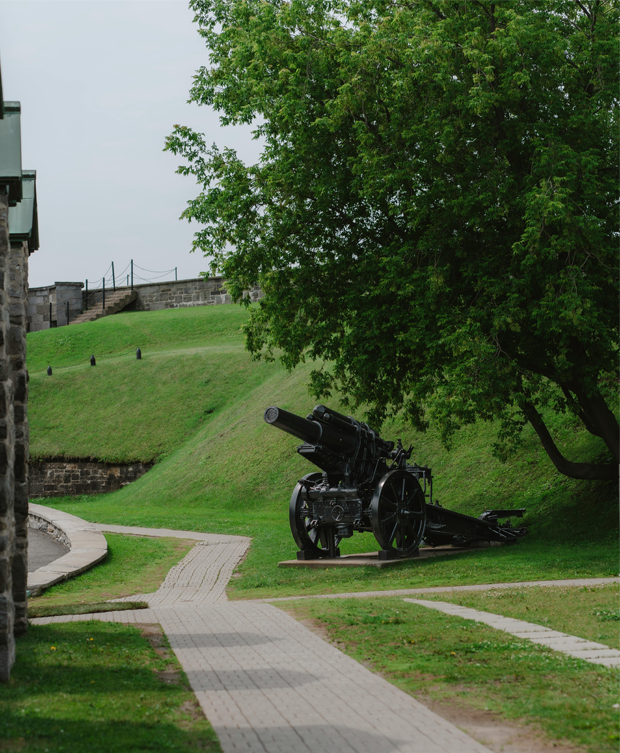 Canon historique installé sur un terrain gazonné entouré d’arbres et de fortifications en pierre, avec un sentier pavé au premier plan.
