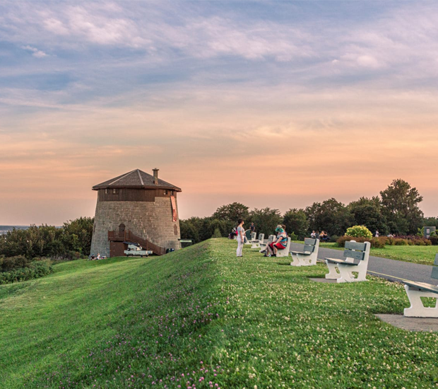 Vue des plaines d’Abraham au coucher du soleil, avec une tour Martello, des visiteurs assis sur des bancs et un vaste espace vert surplombant le paysage.