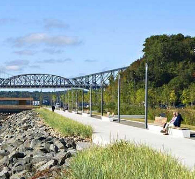 Promenade du parc Samuel-De Champlain longeant le fleuve, avec des visiteurs, des espaces verts et le pont de Québec visible à l’arrière-plan.